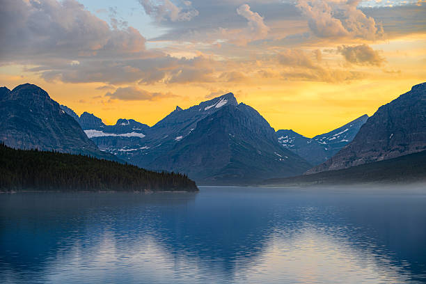 A stunning sunset illuminates the rugged peaks and calm waters of Glacier National Park in Montana. Soft mist floats above the lake as golden light paints the sky, creating a peaceful and awe-inspiring alpine landscape.