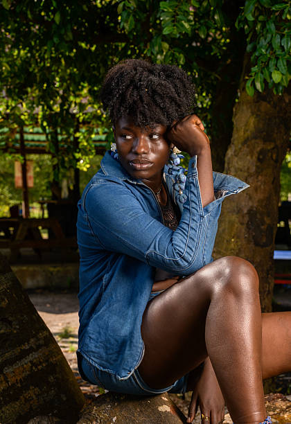 A striking, mid-shot portrait of a young Black woman with up-swept natural curls, seated on a log or tree root outdoors. She wears a full denim jacket, with the collar up and cuffs turned, over a black top, and denim shorts. Her expression is deeply contemplative, looking off-camera while resting her elbow on her knee and her chin in her hand. The image captures a blend of natural style, introspection, and confidence under the dappled light of the trees