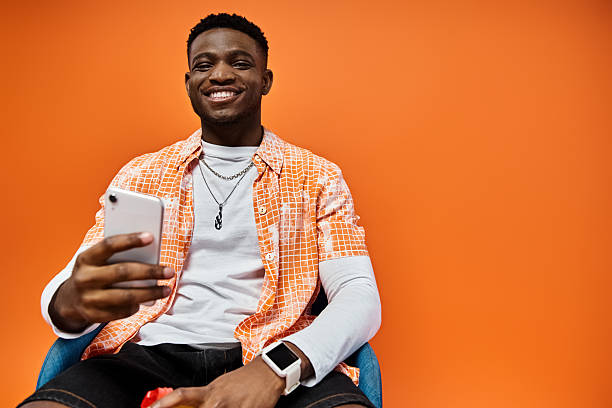istock Young man in stylish attire smiles while holding a phone against a vibrant orange backdrop 2242367237