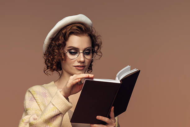 A young woman in glasses and a cozy beret is deeply focused on a book, her soft gaze suggesting a quiet curiosity. She wears a light, patterned sweater, adding to the warm, intellectual vibe of the scene. The soft background tones complement her contemplative expression, creating an inviting, studious atmosphere.