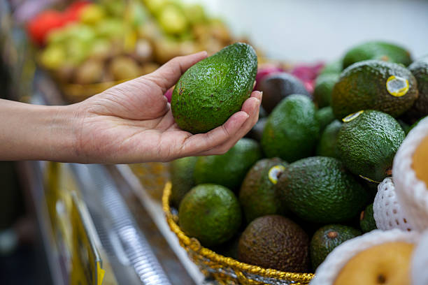 Close-up of a woman holding a ripe avocado while shopping at a local grocery market. Fresh green avocados are displayed in a basket, representing healthy eating, organic produce, and daily lifestyle choices focused on nutrition and sustainability.