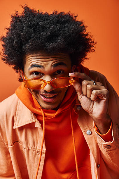 istock Stylish young man expresses joy while posing with vibrant orange glasses and outfit 2242381725