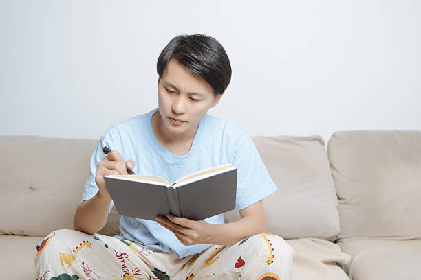 istock Young Asian man sitting on cozy home sofa, holding a notebook and writing while thinking deeply, focused and reflective in bright modern living room. Concept of creativity, personal growth, study, and mindful lifestyle at home. 2242397072