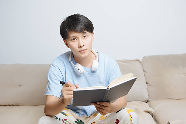istock Young Asian man sitting on cozy home sofa, holding a book and pen while writing, looking at camera with focused and inspired expression in bright modern living room, enjoying personal creativity and solo life. 2242397284