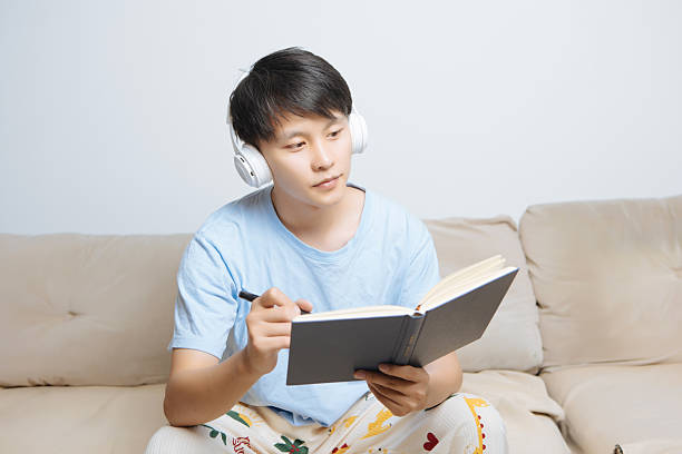 istock Young Asian man sitting on cozy home sofa, holding a book and pen while writing and creating, enjoying solo life and personal leisure in bright modern living room, focused and inspired. 2242397286