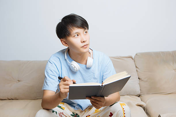 istock Young Asian man sitting on cozy home sofa, holding a book and pen while writing and creating, enjoying solo life and personal leisure in bright modern living room, focused and inspired. 2242397290