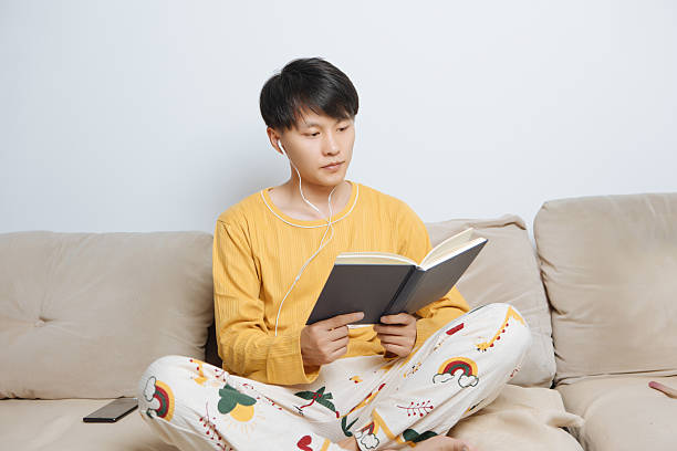 istock Young Asian man sitting on cozy home sofa, reading a book with relaxed and focused expression, enjoying personal leisure and healthy lifestyle in bright modern living room, concept of indoor living and wellness. 2242397430