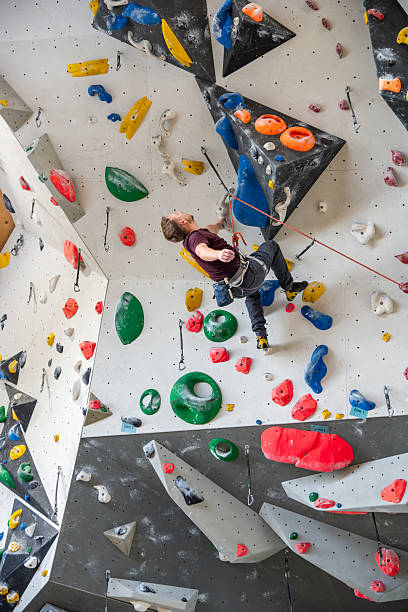 A focused climber wearing a harness climbs a colorful indoor rock wall, gripping holds and navigating routes. The dynamic scene captures determination, athleticism, and the challenging environment of a climbing gym.