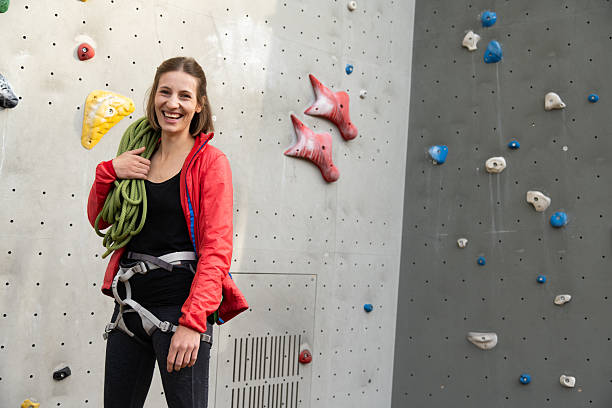 A cheerful woman climber stands beside an indoor climbing wall, holding a coil of rope and wearing a harness. Bright apparel and confident posture convey energy, fitness, and adventure.