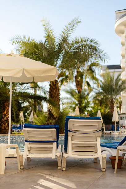 istock Two white lounge chairs with blue cushions sit beside a sparkling pool surrounded by tall palm trees and clear blue skies. A large umbrella provides shade, creating a serene oasis 2242414863