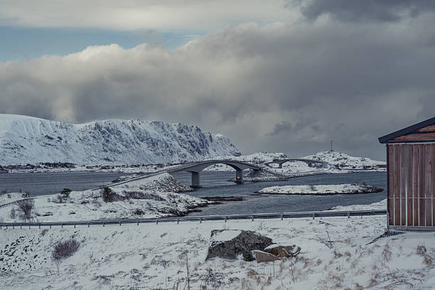 A serene winter landscape showing a snow-covered bridge connecting small islands along the Norwegian coast. The moody sky and rugged terrain reflect the raw beauty of the Arctic environment, where engineering and nature coexist in perfect harmony.