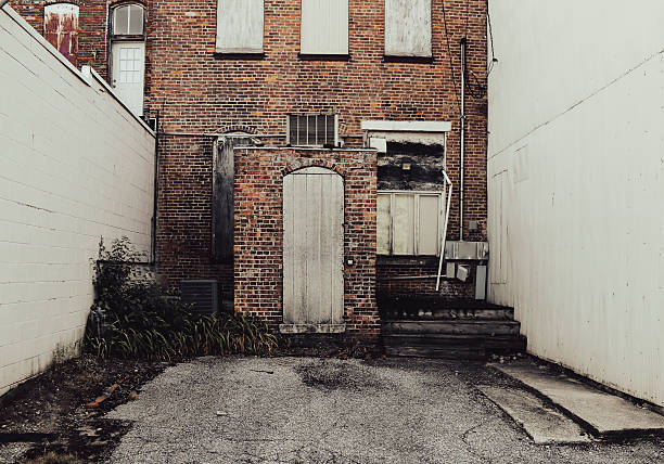 An old brick building with boarded windows and a weathered wooden door sits between two white walls weeds grow by the entrance and cracked pavement leads to the steps