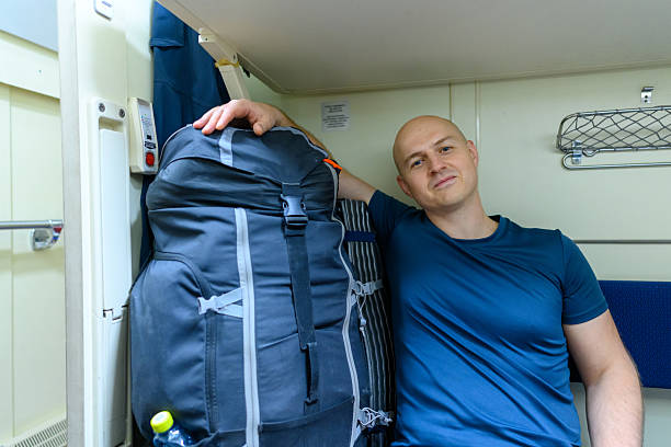 Man sits comfortably in a train cabin, leaning against a large backpack, enjoying a moment of rest on his travels. The space is cozy and inviting.