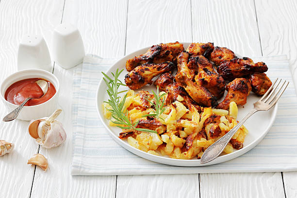 bbq chicken wings with fried potatoes on a plate with rosemary and fork on white wood table with ketchup, horizontal view from above, not AI generated