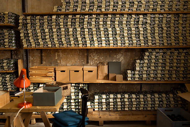 A cluttered storage space filled with neatly stacked coffee on shelves; A wooden workbench with a red lamp, boxes, and bags suggests organized inventory and a busy workspace