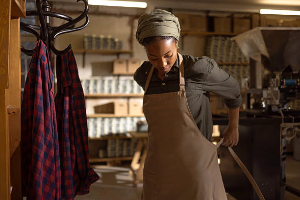A woman wearing a headwrap and apron works in a busy workshop, measuring fabric with a tape, surrounded by shelves and clothing, conveying creativity, craftsmanship, and focused studio work.