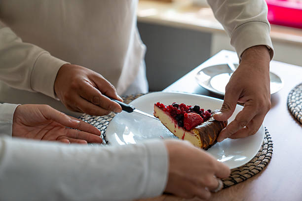 Hands serving a creamy cheesecake slice topped with fresh berries, sharing a sweet homemade dessert between two people at home