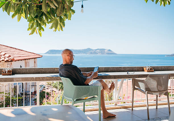 istock Bald man enjoying summer vacation on sunny hotel terrace with seaview surrounded by greenery. He reading last best seller e-book on digital reader device, enjoying leisure time during summer vacation. 2242461723