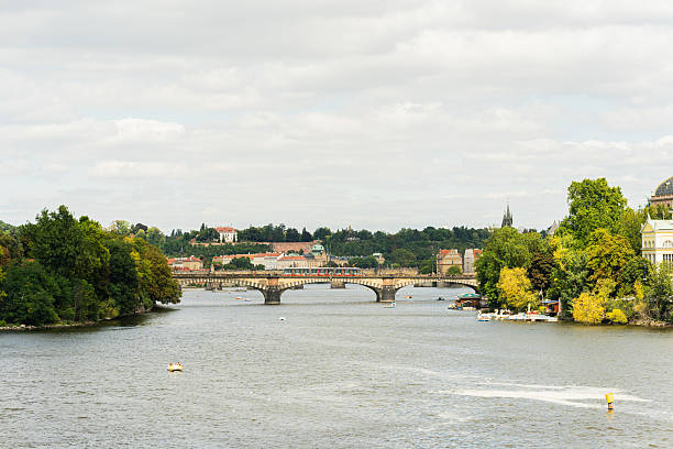 PRAGUE,CZECH REPUBLIC, 10.08.2025: Wide river with boats, lush green trees, and a historic stone bridge in Prague under a cloudy sky. For travel, tourism, architecture and European cityscape themes.