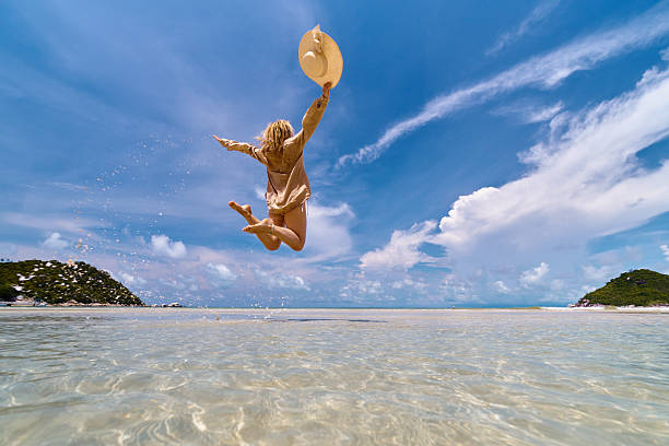 Woman jumping from shallow ocean water with hat against blue sky.