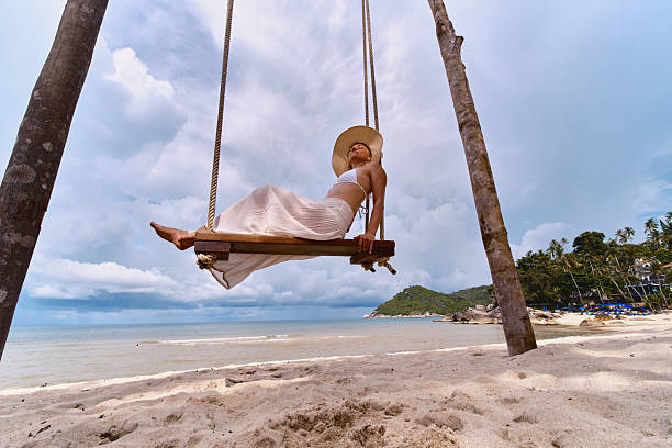Woman wearing a hat and light clothing swinging on a wooden swing at the beach.
