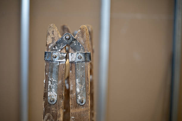 A detailed close-up of a weathered wooden ladder hinge with traces of paint, set against a blurred workshop background. The worn metal and textured wood convey a sense of craftsmanship, labor, and time. The soft focus enhances the rustic and authentic atmosphere of the scene.