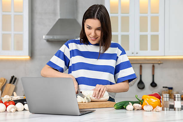 Young woman cooking while watching online course via laptop in kitchen