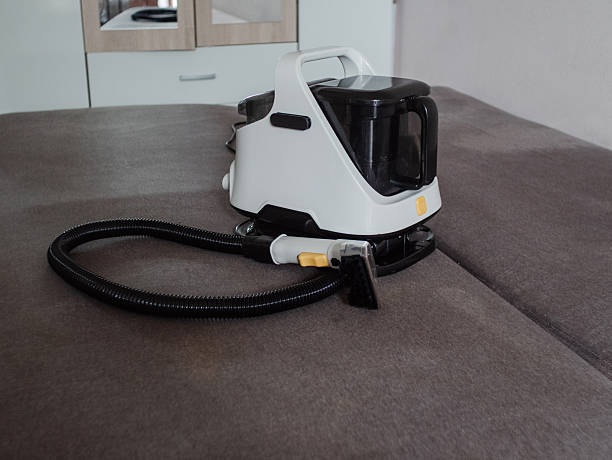 istock A modern white vacuum cleaner with a black container and hose placed on a gray sofa. The background features light-colored furniture and a clean interior. 2242490322