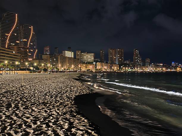 istock foamy shoreline at night, waves softly lapping illuminated shoreline with city backdrop at night 2242490902