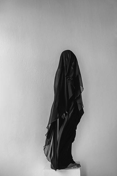 istock Silhouetted figure in a black veil stands on a pedestal against a plain wall 2242508735