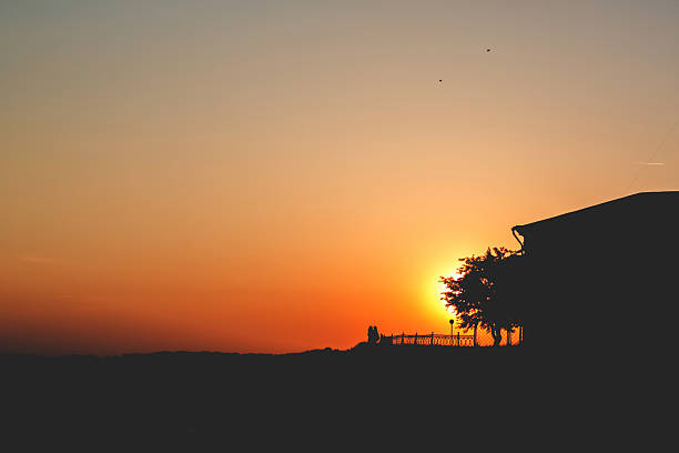 Sunset. Two people stand on a hill with a tree and a house Sunset