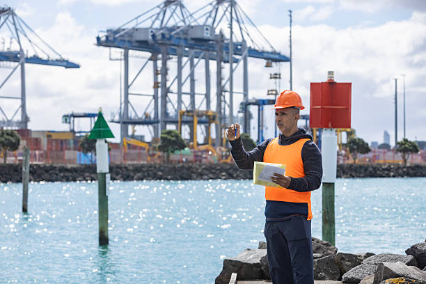 Dubrovnik. Croatia - October 23, 2025: Rear view of a dock worker wearing a yellow hard hat and orange vest. The man is on a concrete pier next to a bollard and uses a red pole to reach the white hull