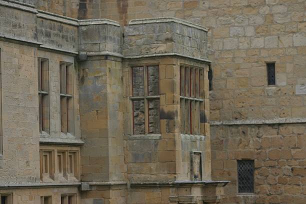 Weathered stone walls and tall mullioned windows form an aged castle facade, showing subtle textures, muted colours, and historic charm in a quiet, timeless architectural scene. Capturing old grandeur.