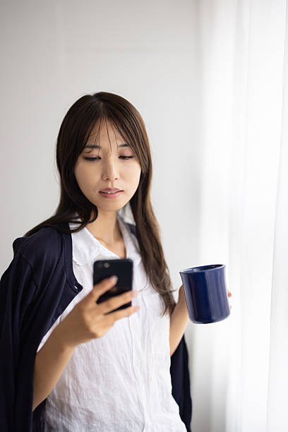Young Woman Using Smartphone While Holding a Mug by the Window