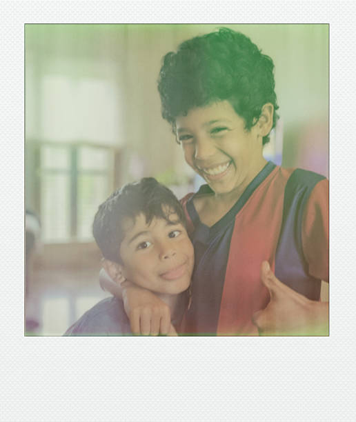 A younger boy playfully sticks his tongue out while being hugged by his smiling older brother in a cozy indoor setting.