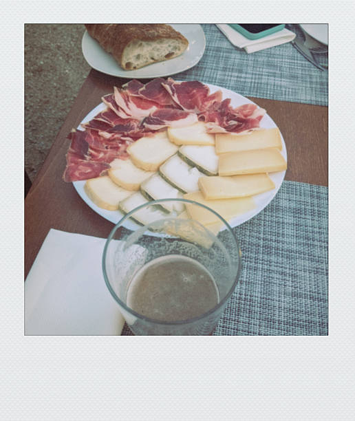 A rustic plate of sliced jamón, assorted cheeses, and crusty bread is served with a glass of beer, ready for a casual meal.