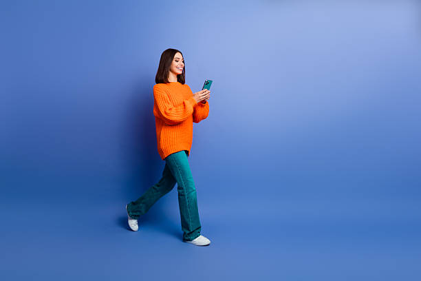 istock Young woman in orange sweater using smartphone while walking on blue background showcasing modern and casual style 2242533809