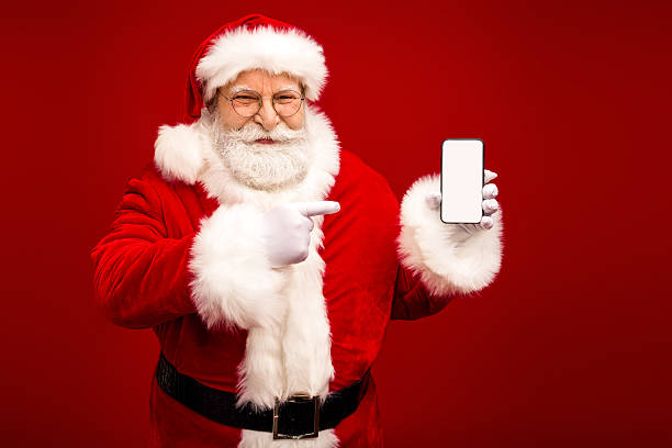istock Santa Claus shows a blank smartphone screen and points at it against a bright red background for a Christmas themed stock image 2242534954