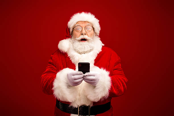 istock Santa Claus in a red suit with white fur trim holds a smartphone against a solid red background ready for Christmas shopping 2242534969