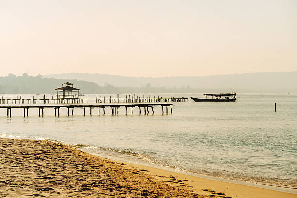 A calm Koh Rong island beach with a long wooden pier extending over gentle waters and a small boat gliding nearby. Soft haze creates a serene, tropical coastal mood.