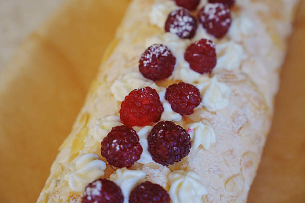A creamy dessert roll features whipped cream and fresh raspberries on top. The sweet treat is beautifully arranged and presented on a smooth wooden surface, perfect for any celebration.
