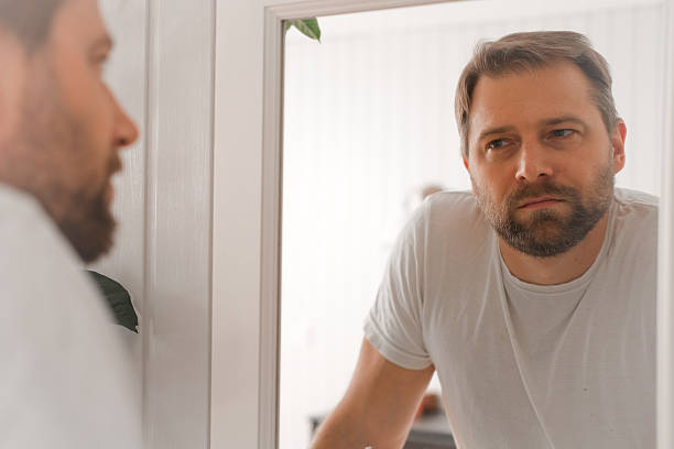 istock Confident handsome bearded man looking himself at the mirror. Pensive guy gazing thoughtfully. Captures introspection, grooming, hair care. Suitable for male health, lifestyle or personal care content 2242552392