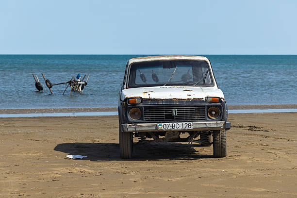 Buzovna, Azerbaijan - March 29, 2025: A weathered white vintage 1987 Lada Niva car sits on the sandy shore as blue waves meet the horizon