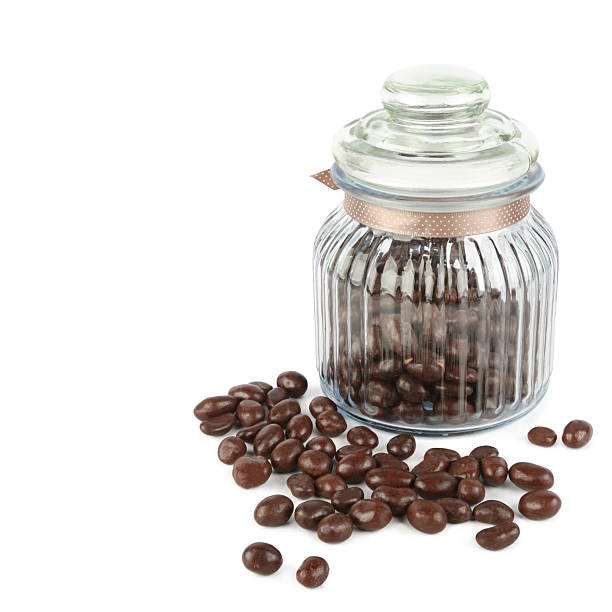 A clear, ribbed glass jar filled with chocolate-coated candies (likely coffee beans or nuts) stands next to a small pile of spilled sweets, all isolated on white. The image offers excellent copy space and evokes themes of confectionery and indulgence.