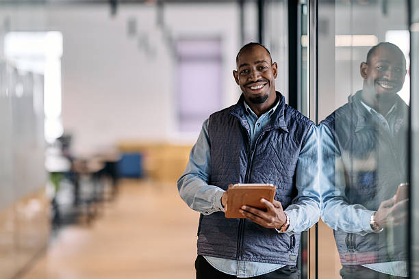 istock Confident african american businessman standing in a modern office hallway, smiling while using a digital tablet. Reflection in glass wall symbolizes technology, success, and innovation in business. 2242560726