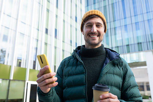 istock Smiling young man using smartphone and holding coffee outside office building 2242570659