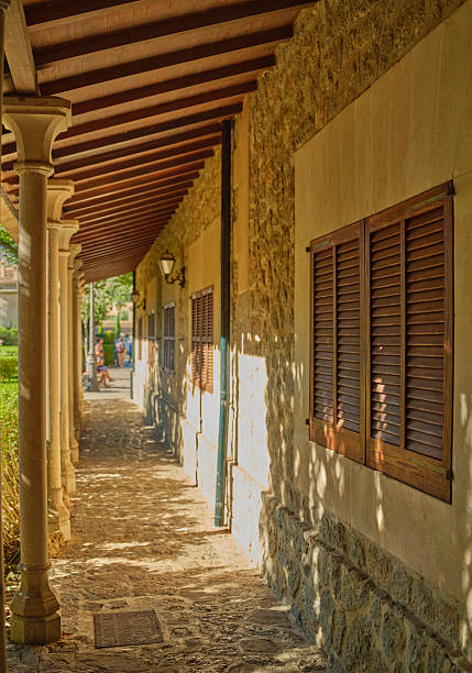 A peaceful stone pathway under a shaded archway leads alongside a building with wooden shutters on the island of Majorca, Spain. The light filters through surrounding trees, creating beautiful patterns.