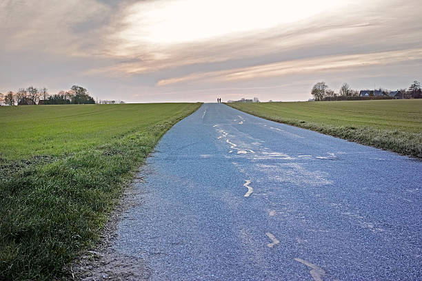 A quiet countryside scene with a paved road stretching into the horizon, bordered by green fields. The setting sun casts soft hues across the sky, creating a peaceful and open atmosphere.