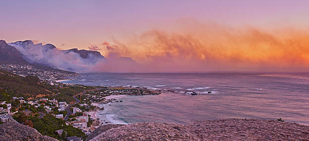 A sweeping panorama of the Cape Town coast at sunset. The scene blends warm orange-pink skies with misty mountains, a tranquil ocean, and a quiet coastal town tucked along the Western Cape. This image evokes serenity, travel, and the natural beauty of South Africaâs coastline.