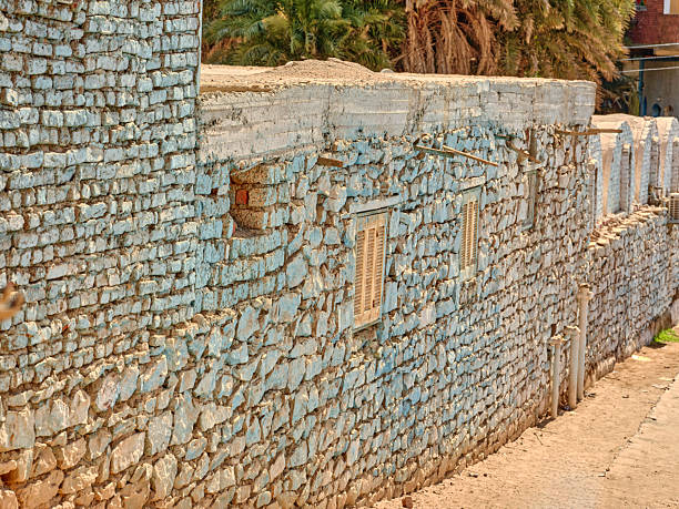 The image showcases a light blue-painted stone wall, featuring wooden-framed windows, in a quaint Egyptian village setting. Palms and a sunny backdrop emphasize the traditional and rustic architectural style.